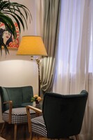 Una stanza con una poltrona verde, una sedia decorata, una lampada, una pianta e un quadro appeso al muro.