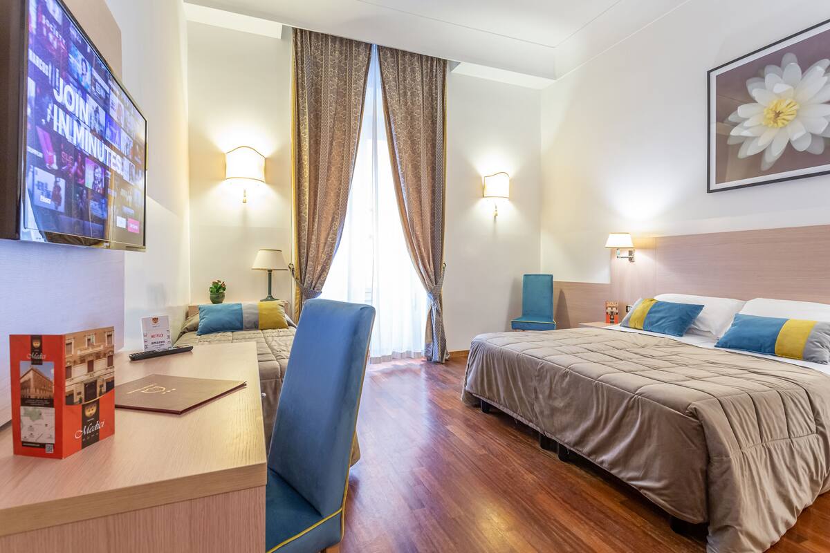 A hotel room with a wooden floor, a desk with a laptop, a chair, a bed with blue and yellow pillows, and a large flower painting on the wall.