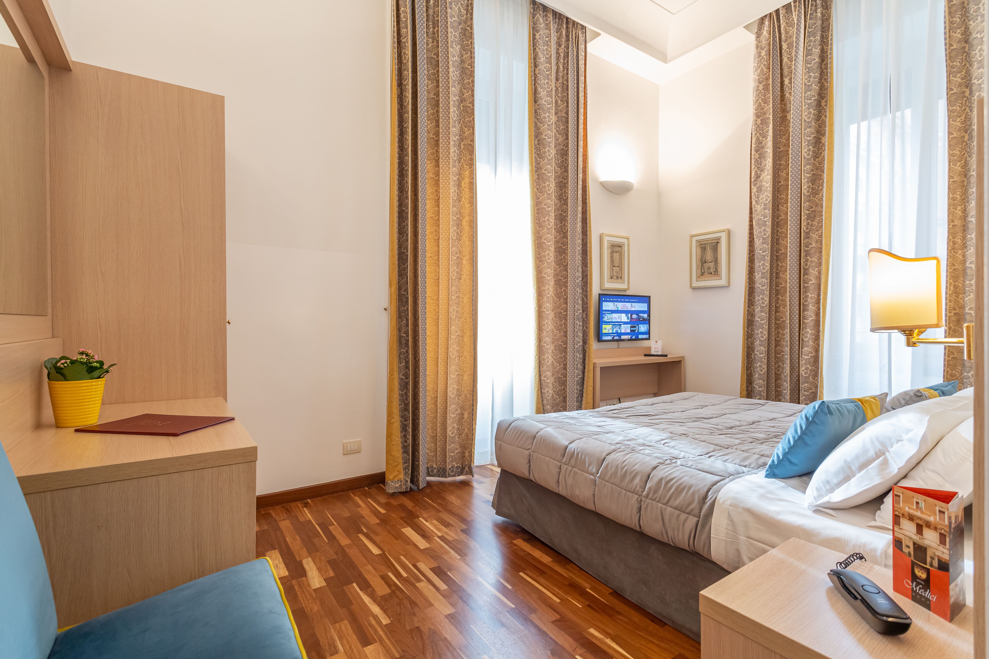 A hotel room with a bed, bedside table, TV, lamp, and a small desk with a plant.
