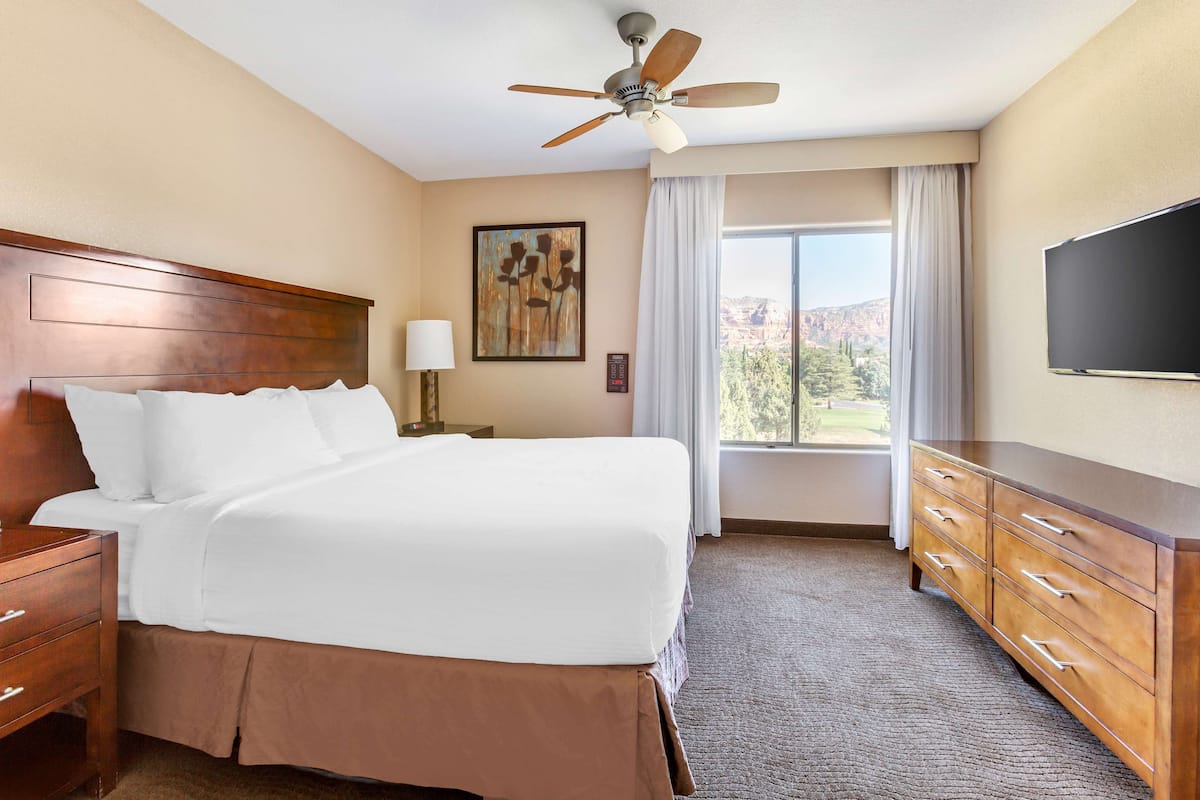 A hotel room with a large bed, a wooden headboard, a ceiling fan, a TV, a nightstand, a lamp, a dresser, and a window with curtains.