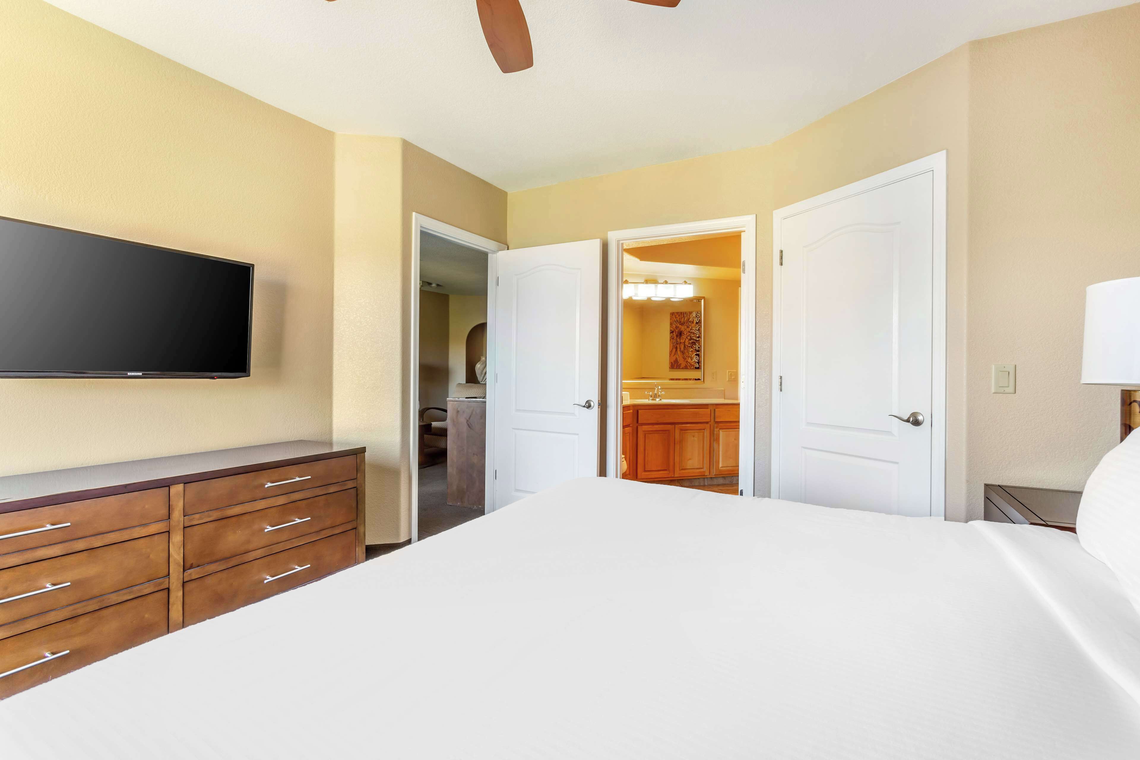 A hotel room with a bed, a TV mounted on the wall, a wooden dresser, and a bathroom visible through an open door.