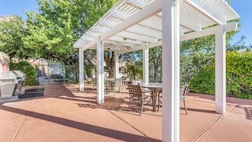 A covered patio area with tables and chairs, surrounded by greenery and a barbecue grill.