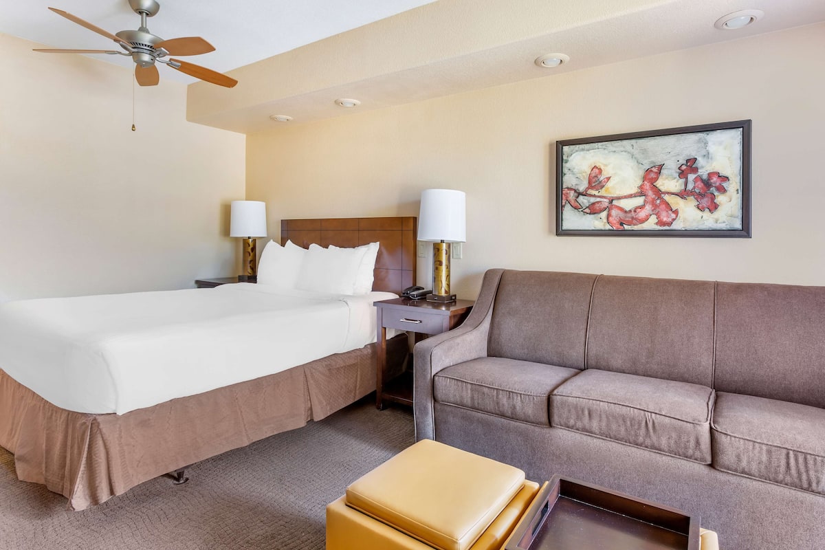A hotel room with two beds, a sofa, a ceiling fan, a nightstand, lamps, and a framed artwork on the wall.
