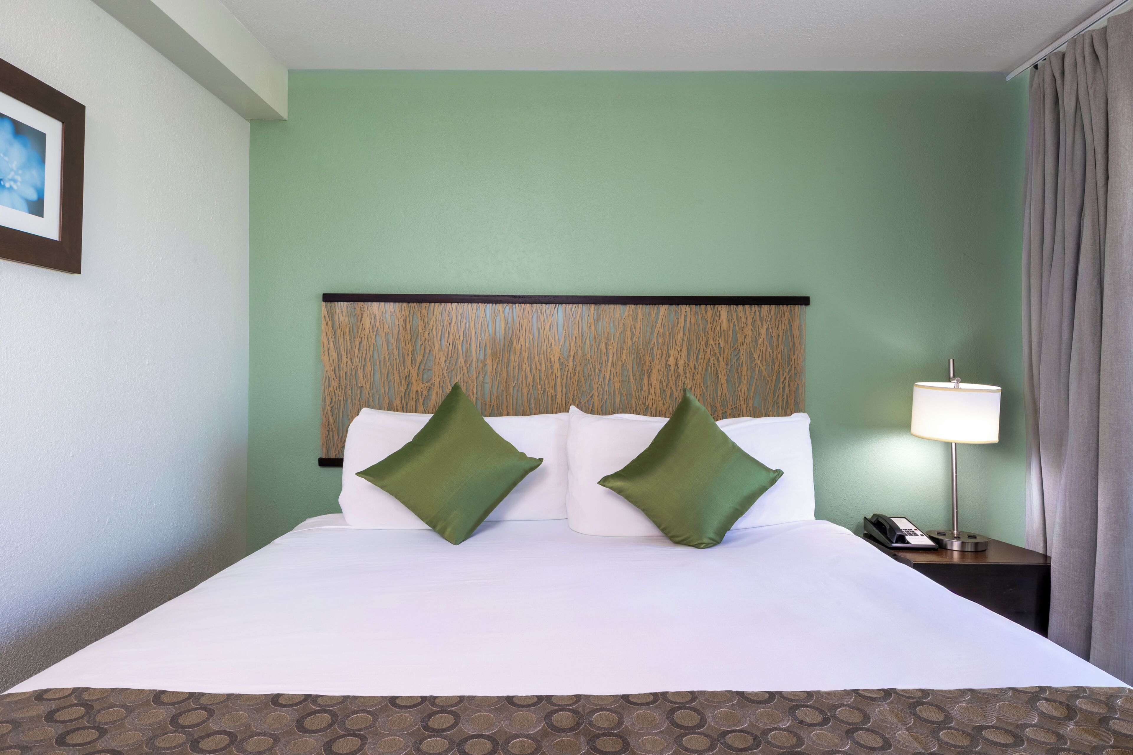 A hotel room with a bed, two green pillows, a nightstand with a lamp, and a painting on the wall.