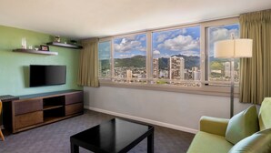 A living room with a TV, sofa, coffee table, and a large window with a city view. - Skyline Island Colony Hotel (Honolulu)