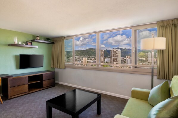 A living room with a TV, sofa, coffee table, and a large window with a city view. - Skyline Island Colony Hotel (Honolulu)