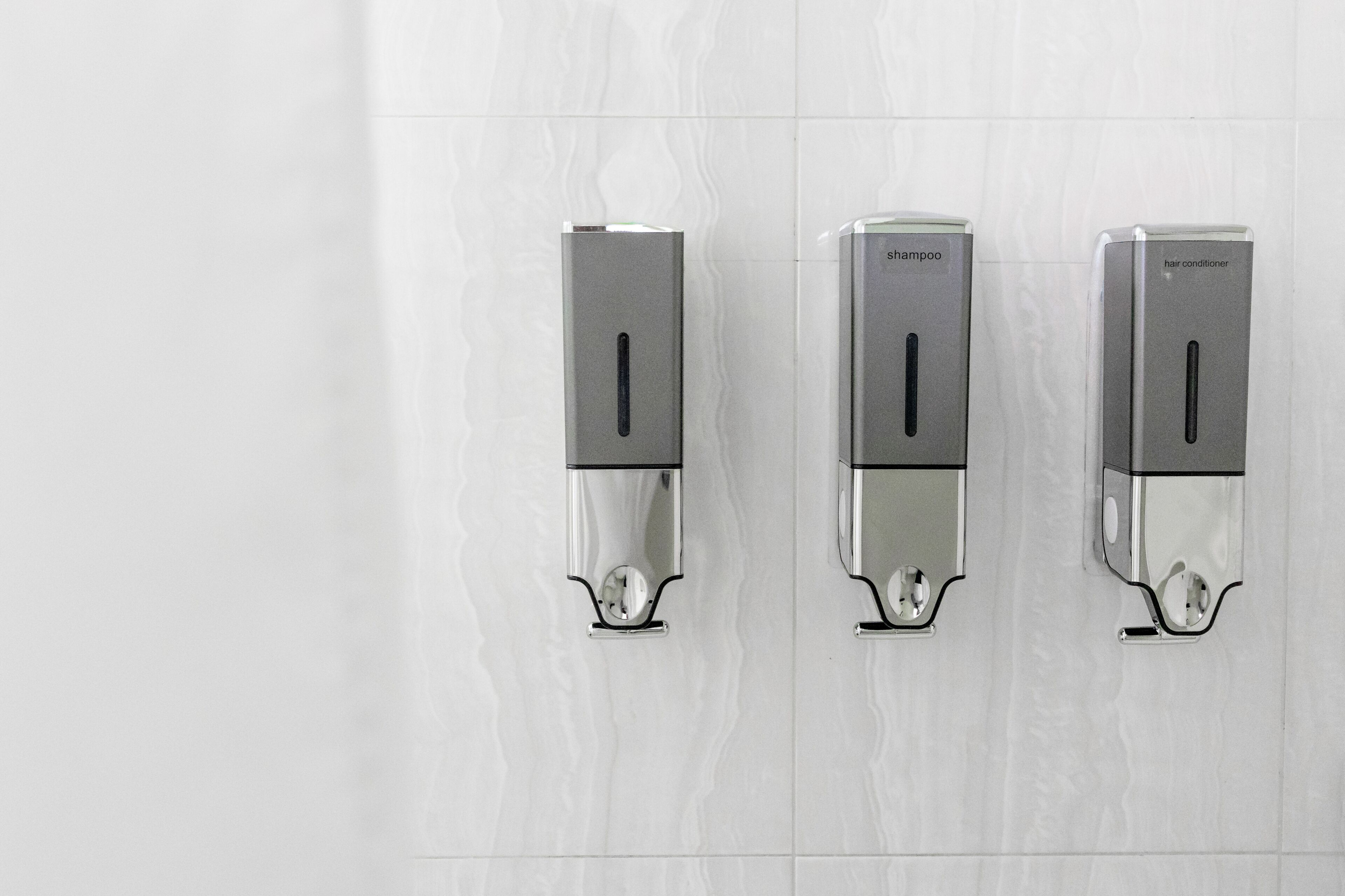 Dans une salle de bain aux murs recouverts de carreaux blancs, on trouve trois distributeurs fixés au mur, étiquetés respectivement « shampoing », « shampoing » et « après-shampoing ».