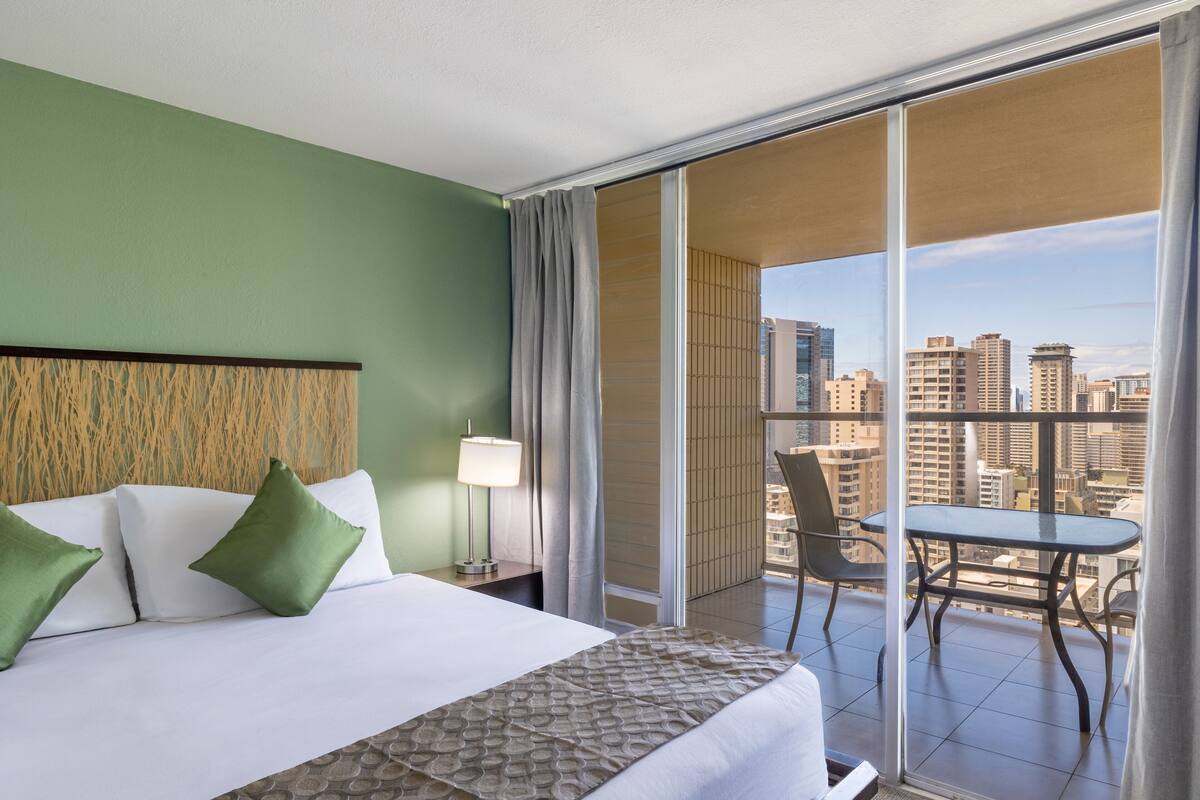A hotel room with a bed, a nightstand, a lamp, a balcony with a table and chairs, and a city view.