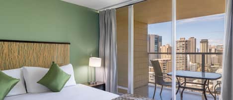 Una habitación de hotel con cama, mesita de noche, lámpara, balcón con mesa y sillas, y vista a la ciudad.