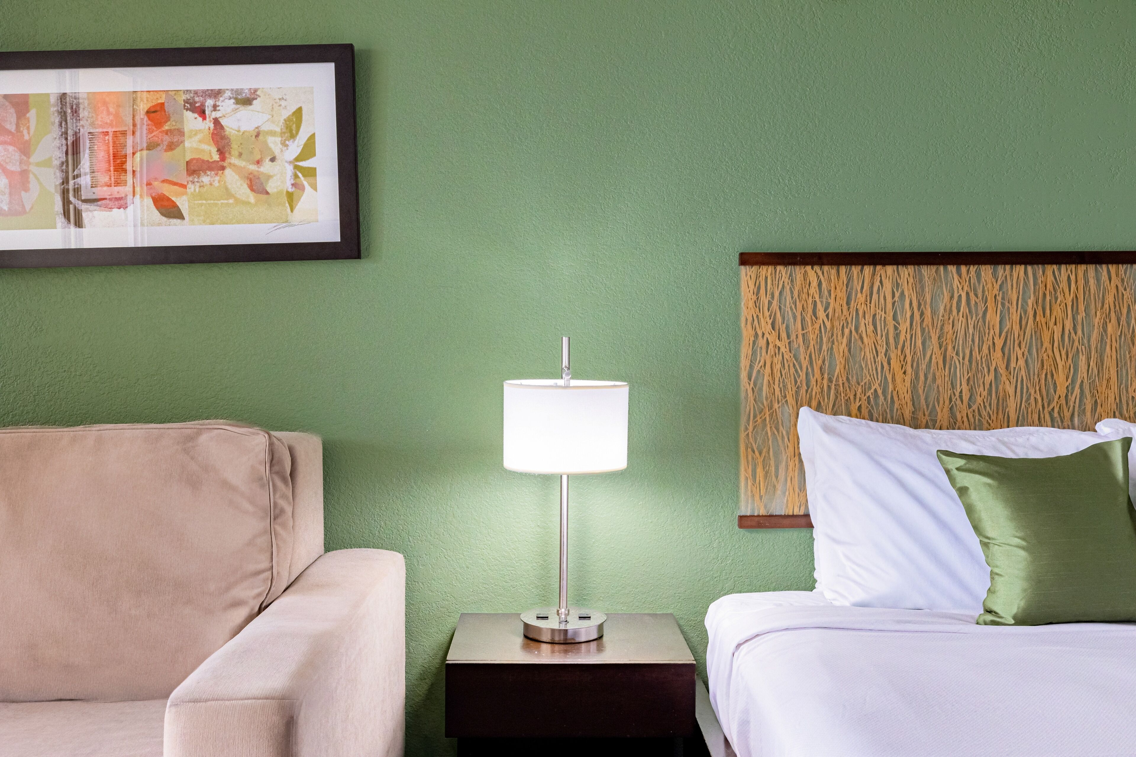 A hotel room with a bed, a nightstand with a lamp, a framed picture on the wall, and a green textured wall.