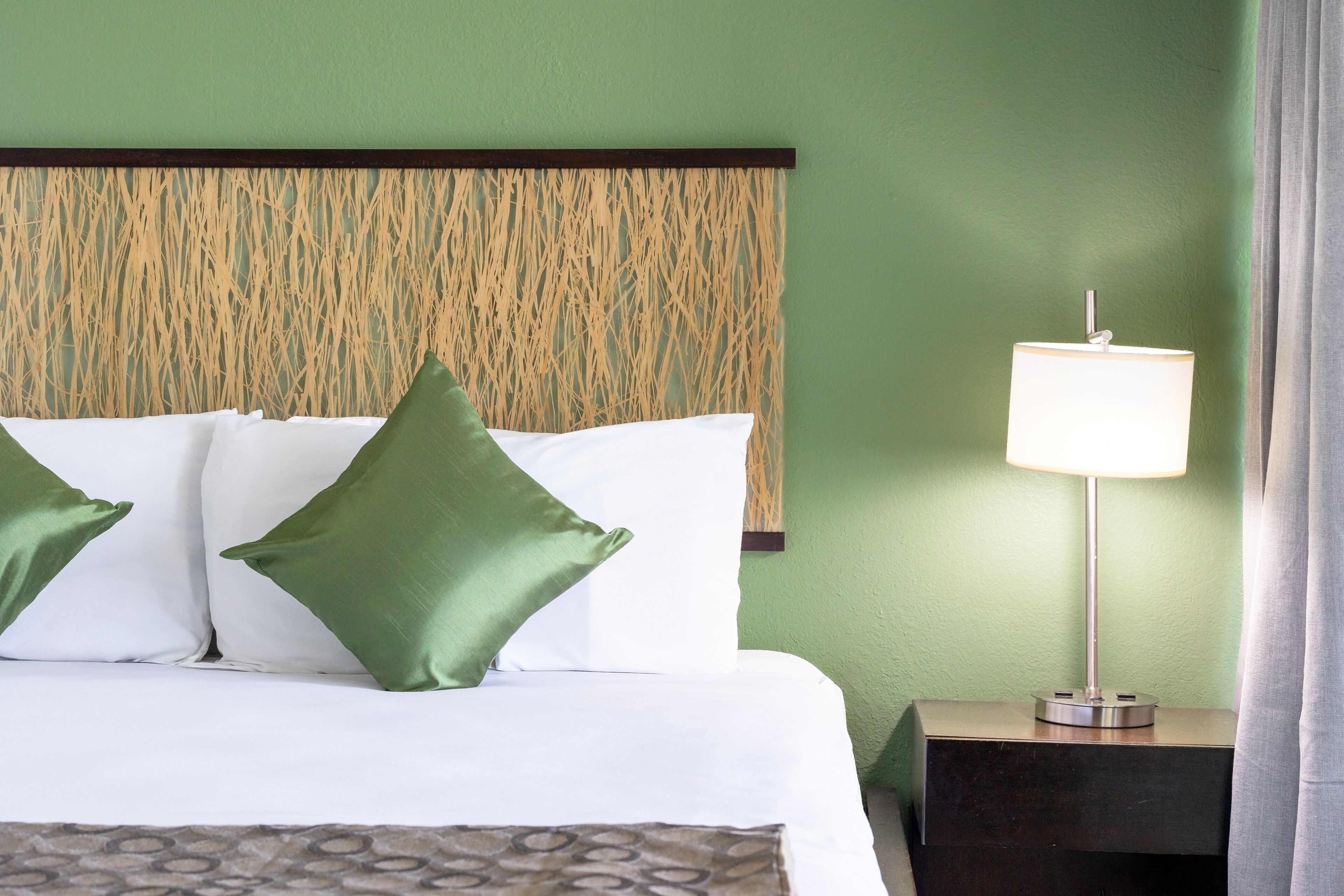 A neatly made bed with green and white pillows, a patterned bedspread, and a bedside lamp on a nightstand.