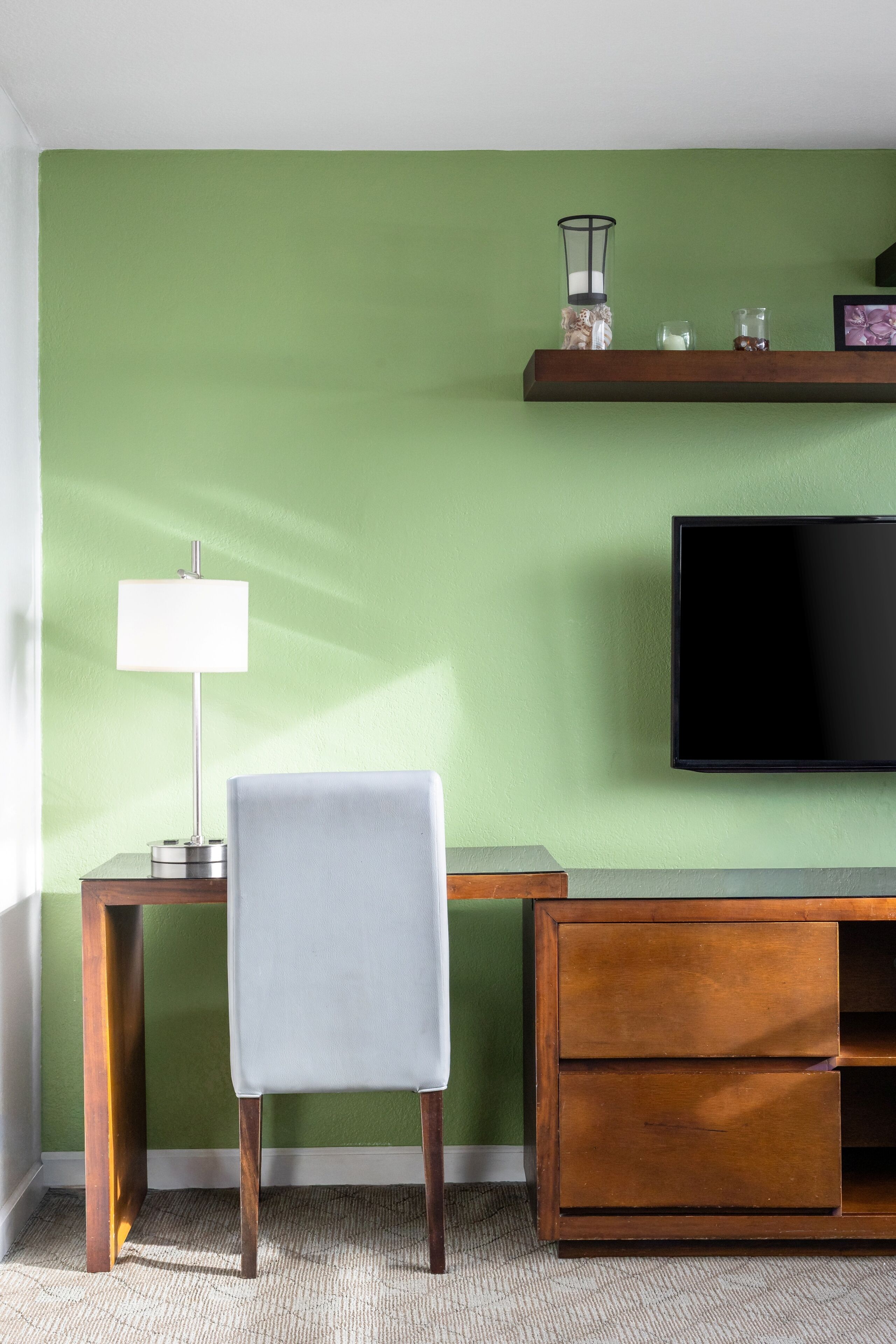 Une pièce avec un mur vert, un téléviseur à écran plat, un bureau avec une lampe, une chaise et un meuble en bois.