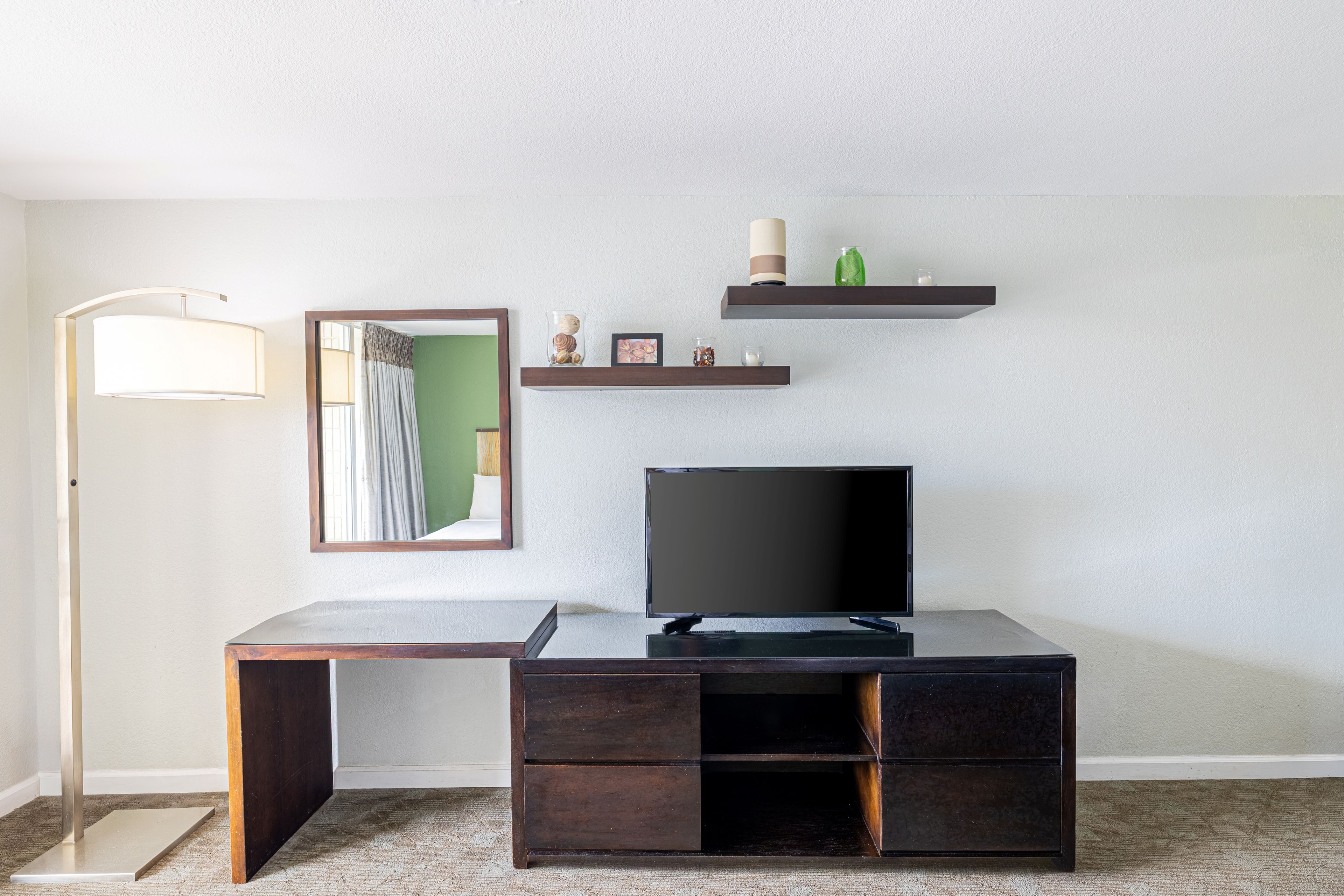 Une chambre moderne avec un téléviseur à écran plat posé sur un bureau en bois foncé, un lampadaire et un miroir accroché au mur.