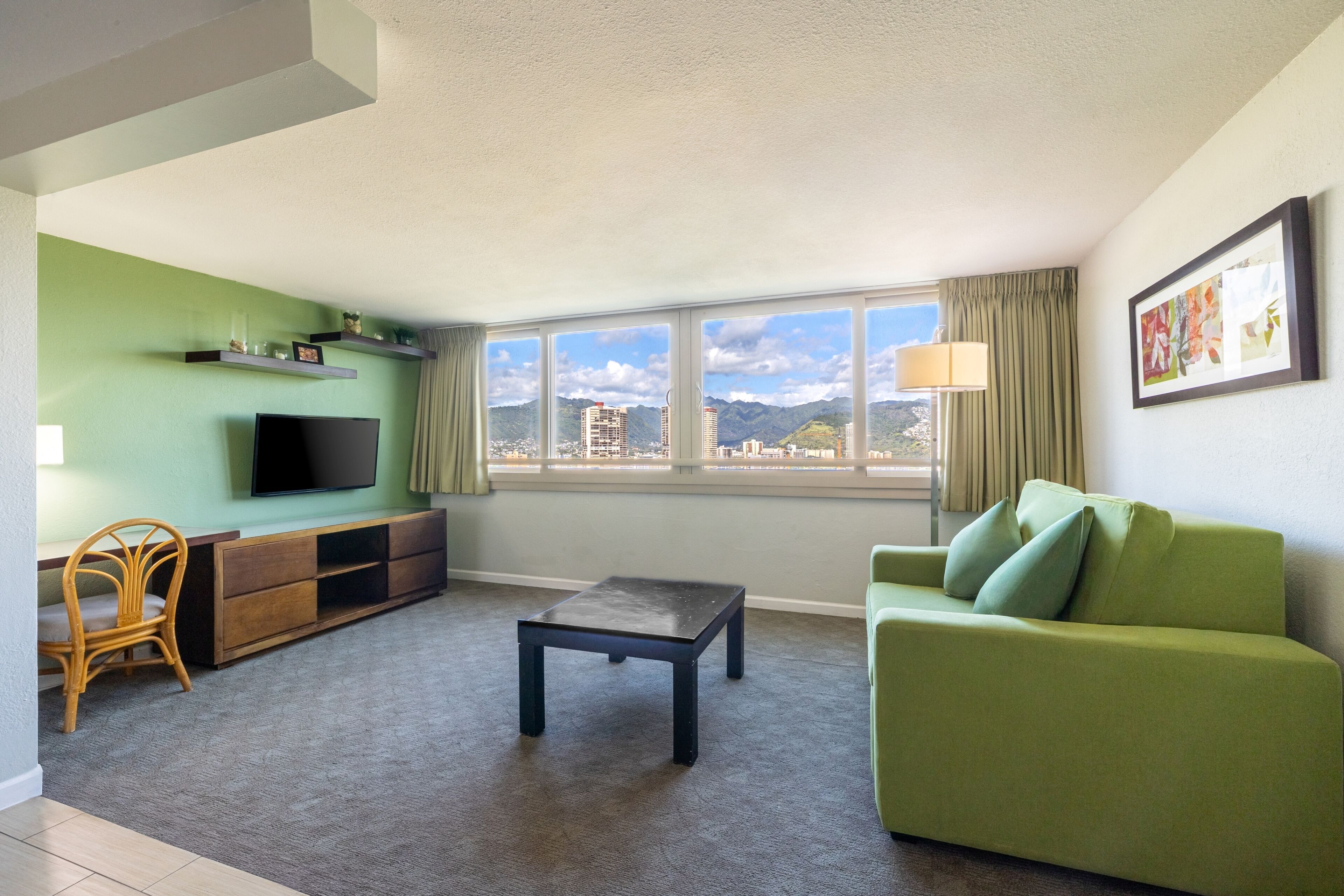 A living room with a green sofa, a TV on a stand, a small table, and a window with a view of buildings and mountains.