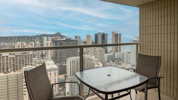 A balcony with a table and chairs, overlooking a cityscape with high-rise buildings and mountains. - Skyline Island Colony Hotel (Honolulu)