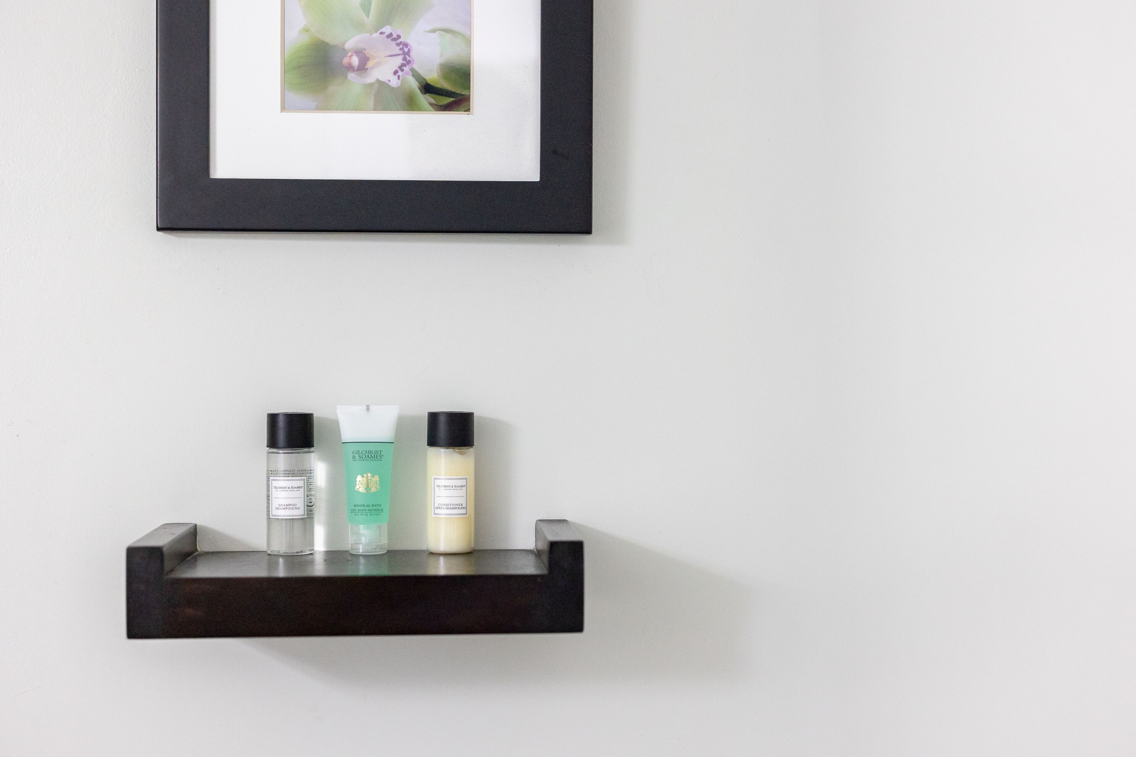 A framed picture of a flower on a wall, a dark wooden shelf with three bottles, and a white wall.
