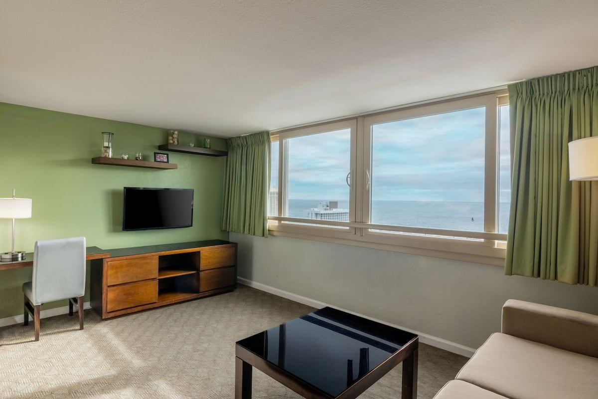 A room with a TV, a desk, a chair, a sofa, and a window with a view of the sea.