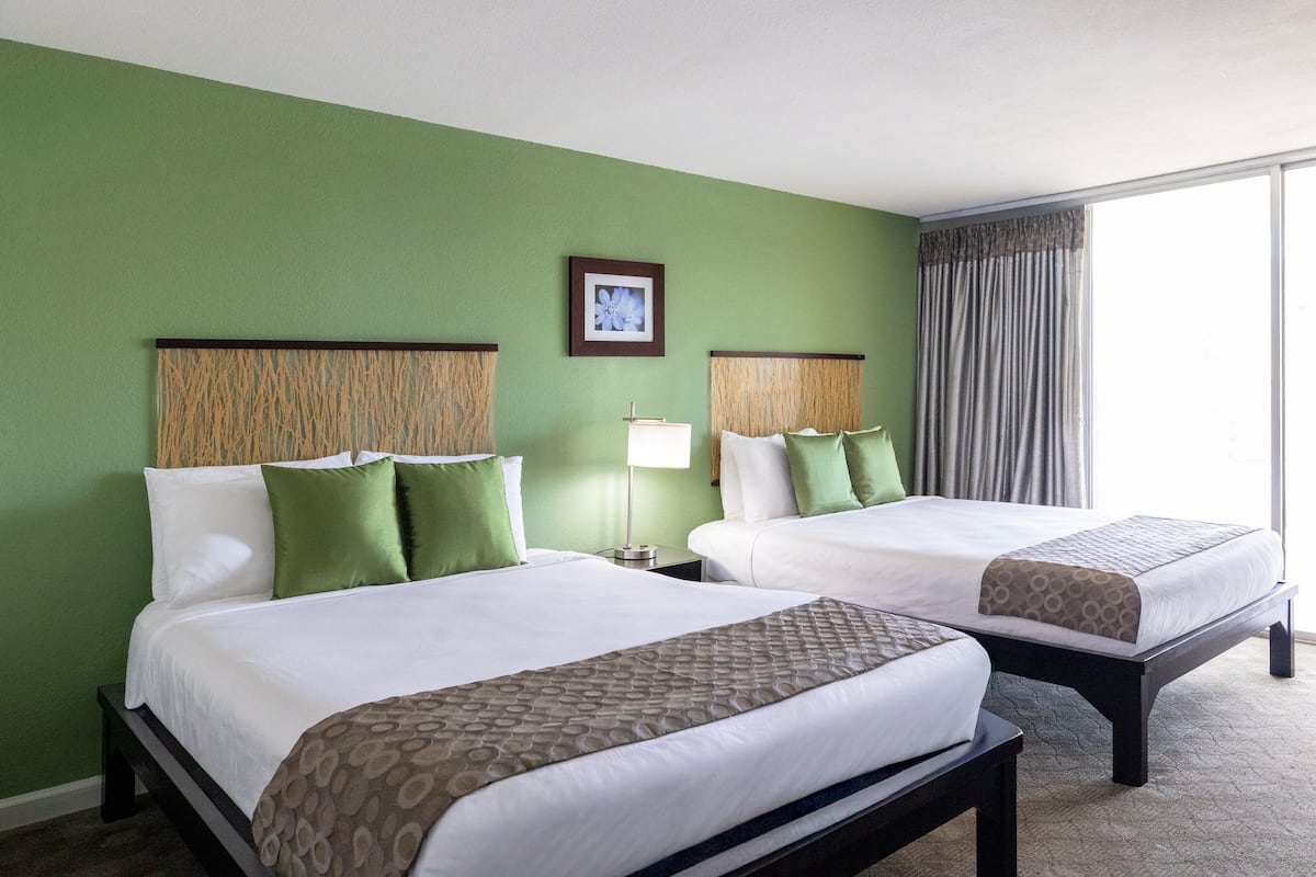 A hotel room with two beds, green walls, a framed picture, a window with curtains, and a bedside table with a lamp.