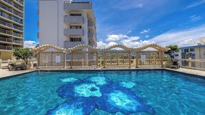A swimming pool with clear blue water, surrounded by a wooden fence and a white building with balconies. - Skyline Island Colony Hotel (Honolulu)