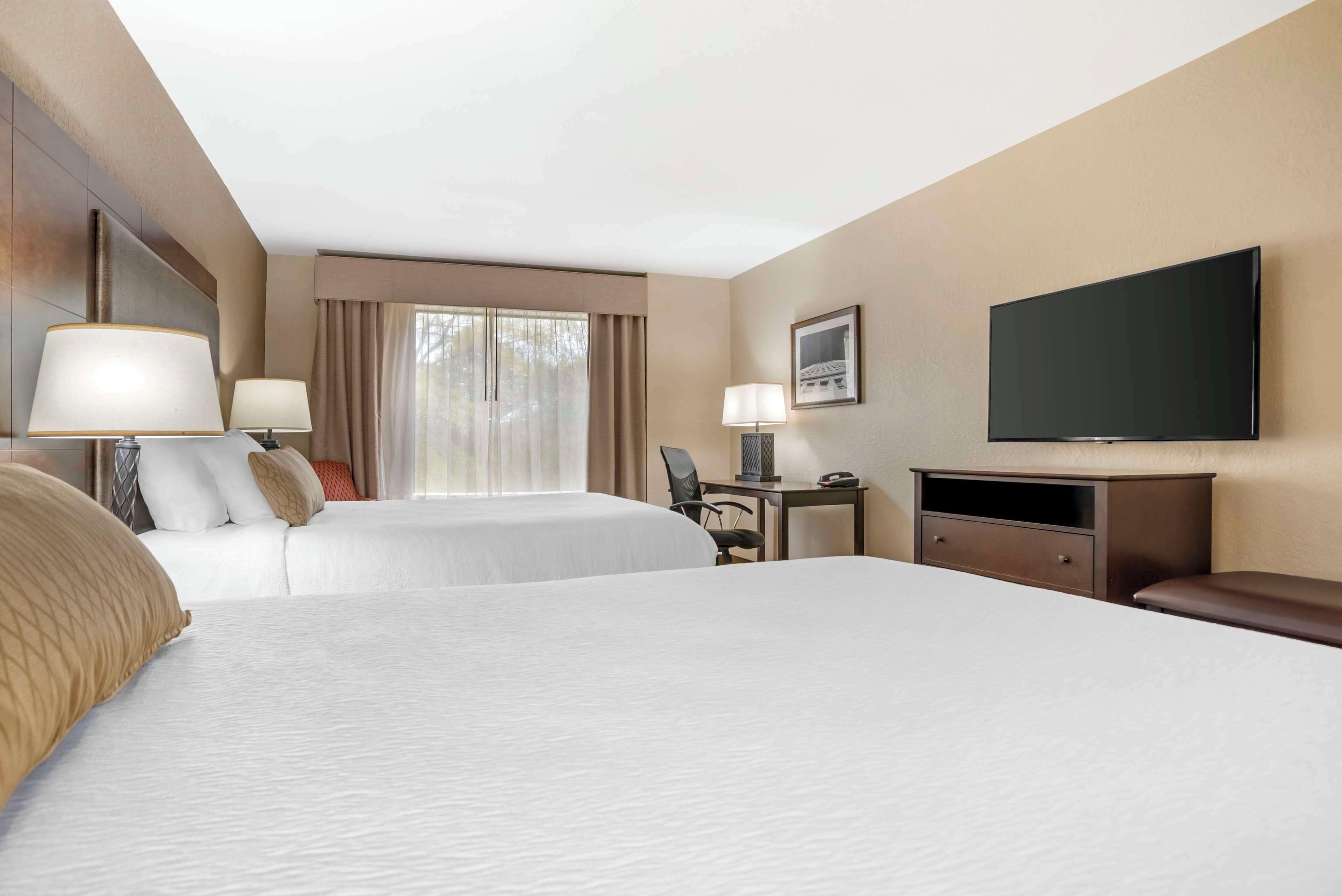 A hotel room with two beds, a desk, a chair, a flat-screen TV, and a window with curtains.