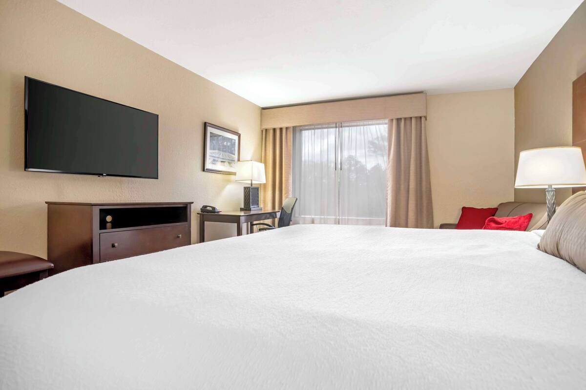 A hotel room with a large bed, a flat-screen TV mounted on the wall, a desk with a lamp, and a window with curtains.