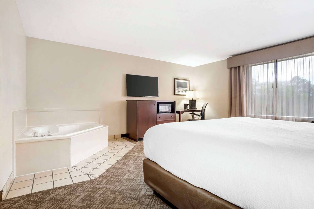 A hotel room with a large bed, a separate bathtub, a desk with a chair, a flat-screen TV, and a window with curtains.