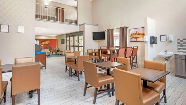 A hotel dining area with multiple tables and chairs, a TV mounted on the wall, and a visible kitchen area with a sink and cabinets. - Best Western Gardendale (Gardendale)