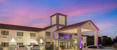 Un hotel con entrada cubierta, varios coches estacionados al frente y cielo despejado.