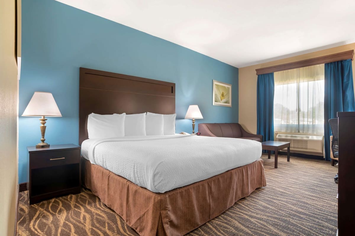 A hotel room with a large bed, two bedside tables with lamps, a chair, a desk, and a large window with curtains.