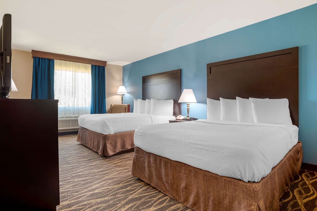 A hotel room with two beds, a desk, a lamp, and a large window with curtains.