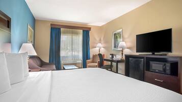 Quarto de hotel com uma cama grande, duas cadeiras, uma escrivaninha com abajur, televisão e minibar.