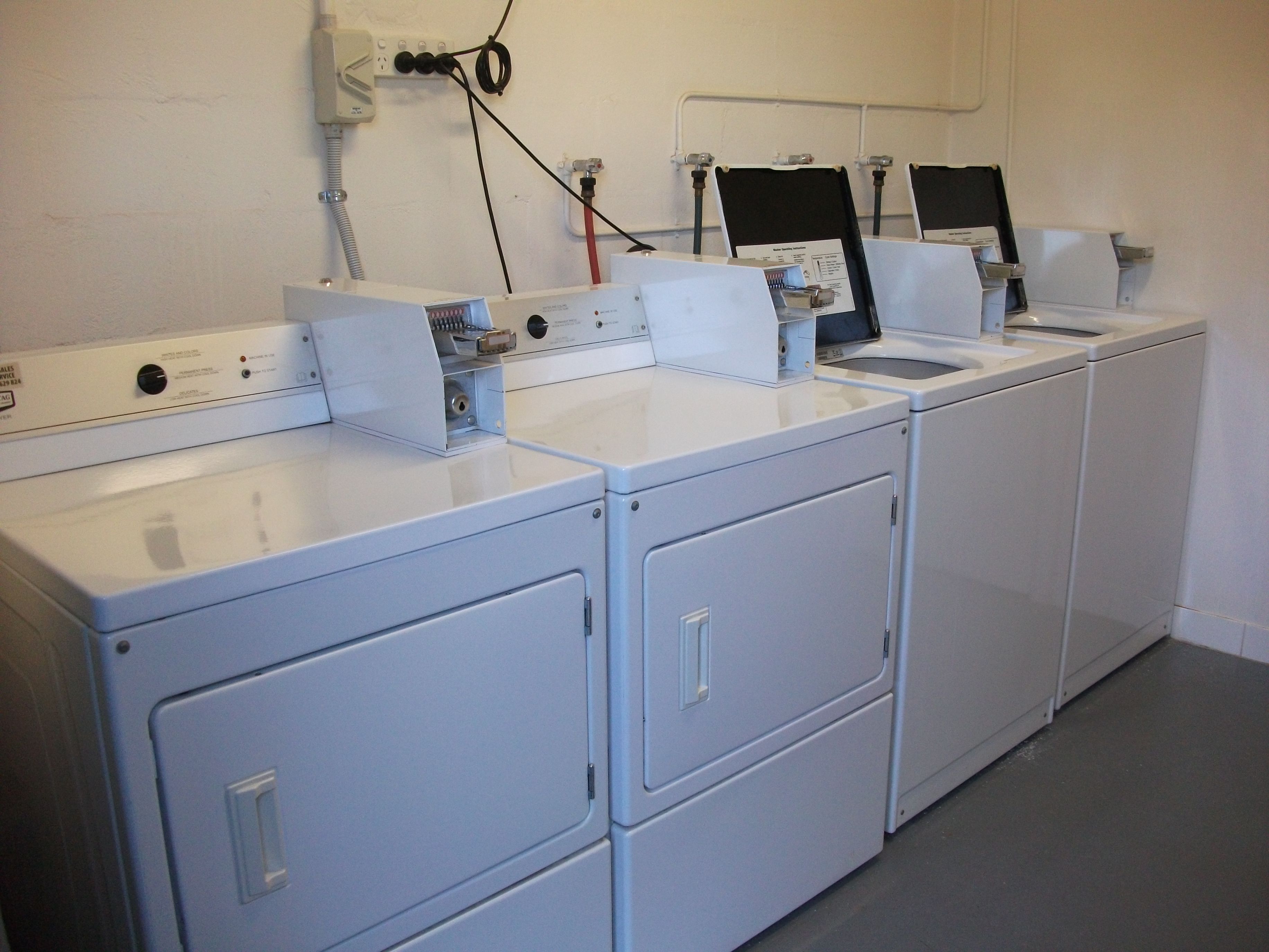 A row of white laundry machines with control panels, set against a plain wall with visible electrical outlets and cables.