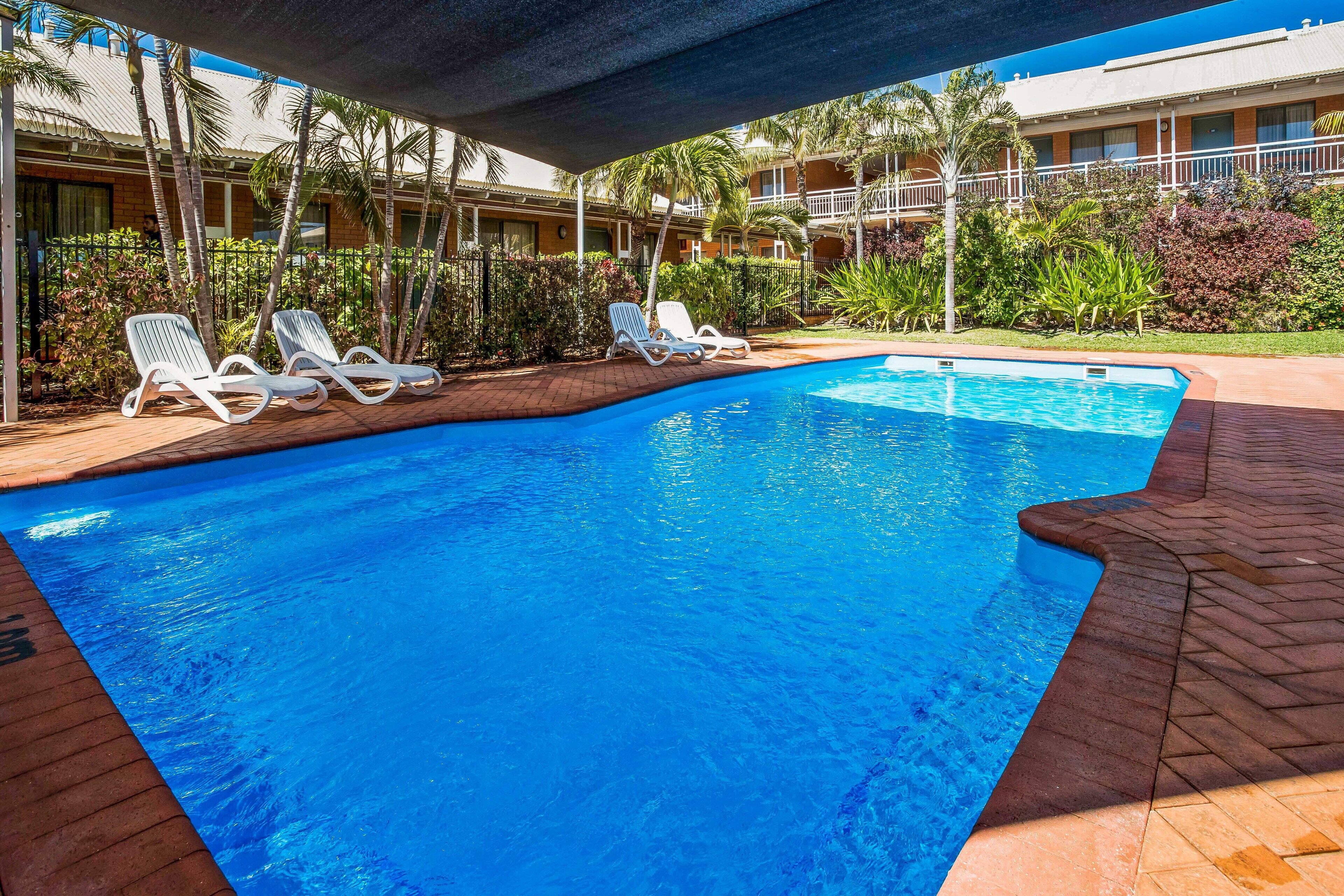 Piscina con sillones de descanso y zona cubierta, rodeada de vegetación y un edificio con balcones.