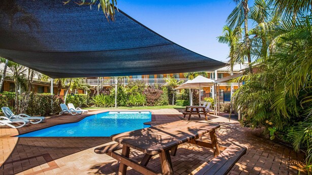 Zona de piscina con mesa y sillas de madera, rodeada de palmeras y un área de descanso cubierta.