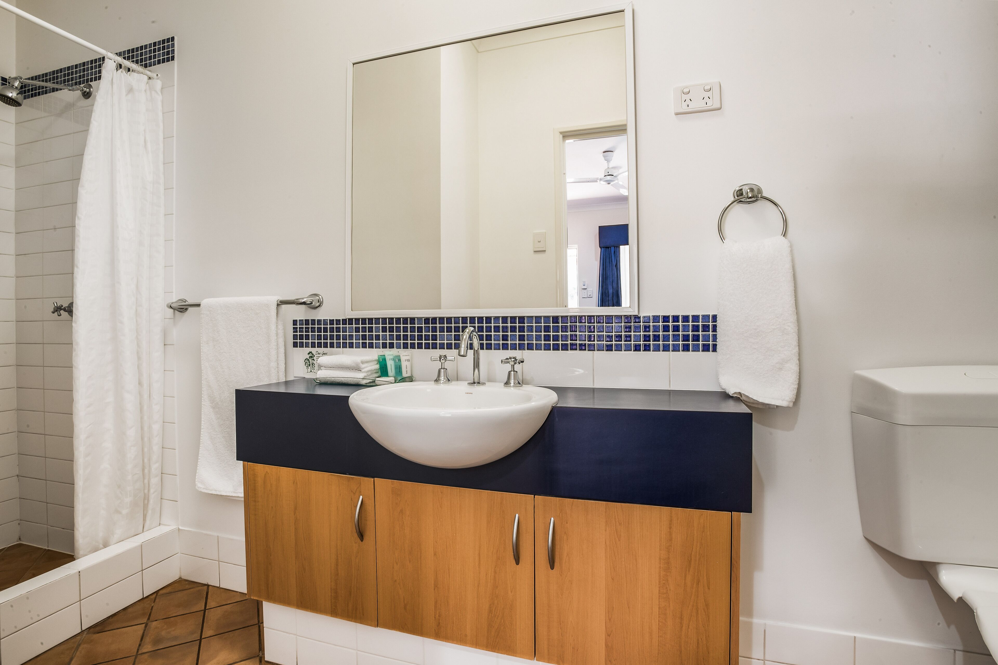 Un baño con lavamanos blanco, encimera oscura, armarios de madera, espejo y ducha con cortina blanca.