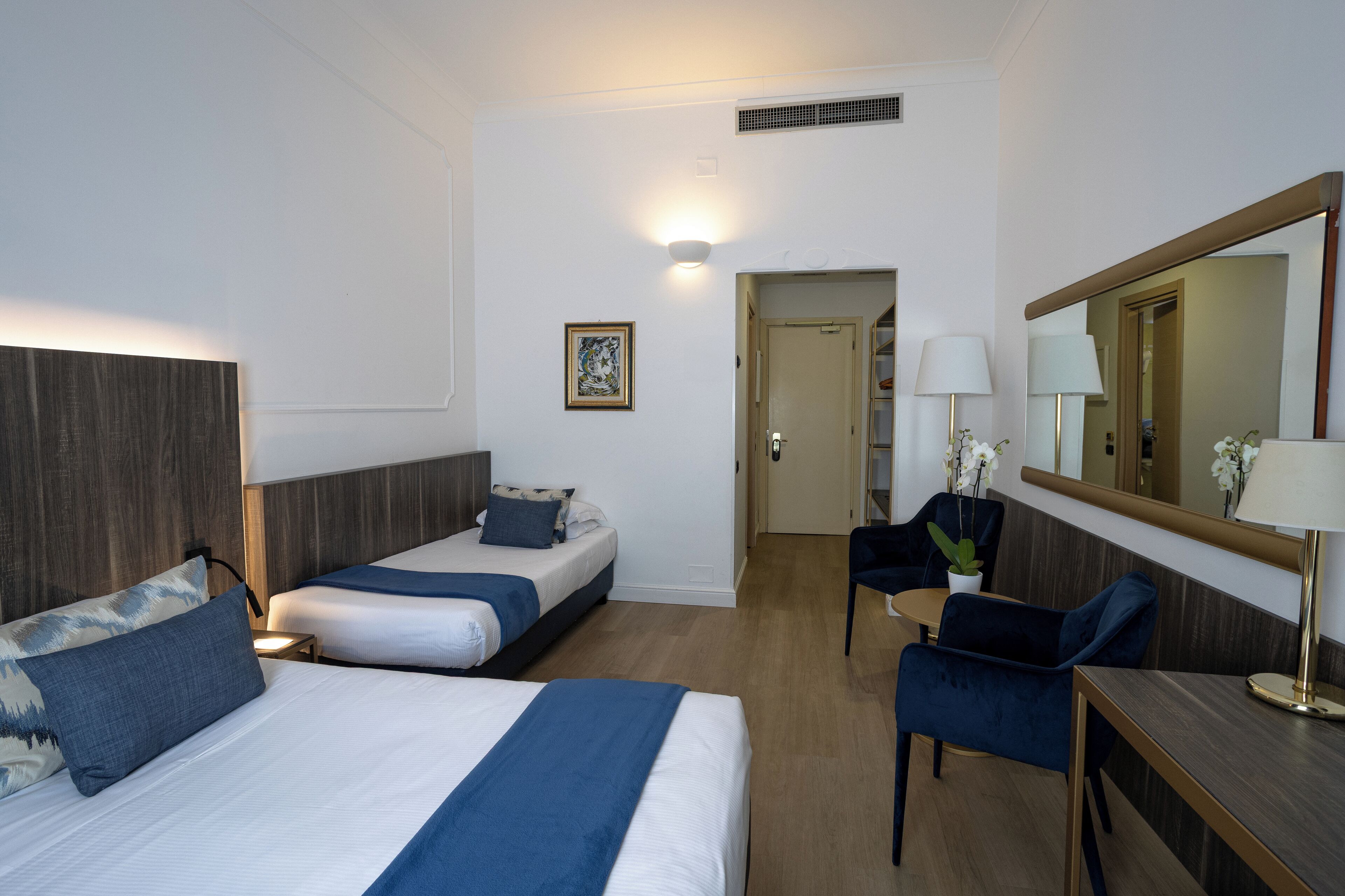 A hotel room with two beds, a desk, a chair, a mirror, and a painting on the wall.