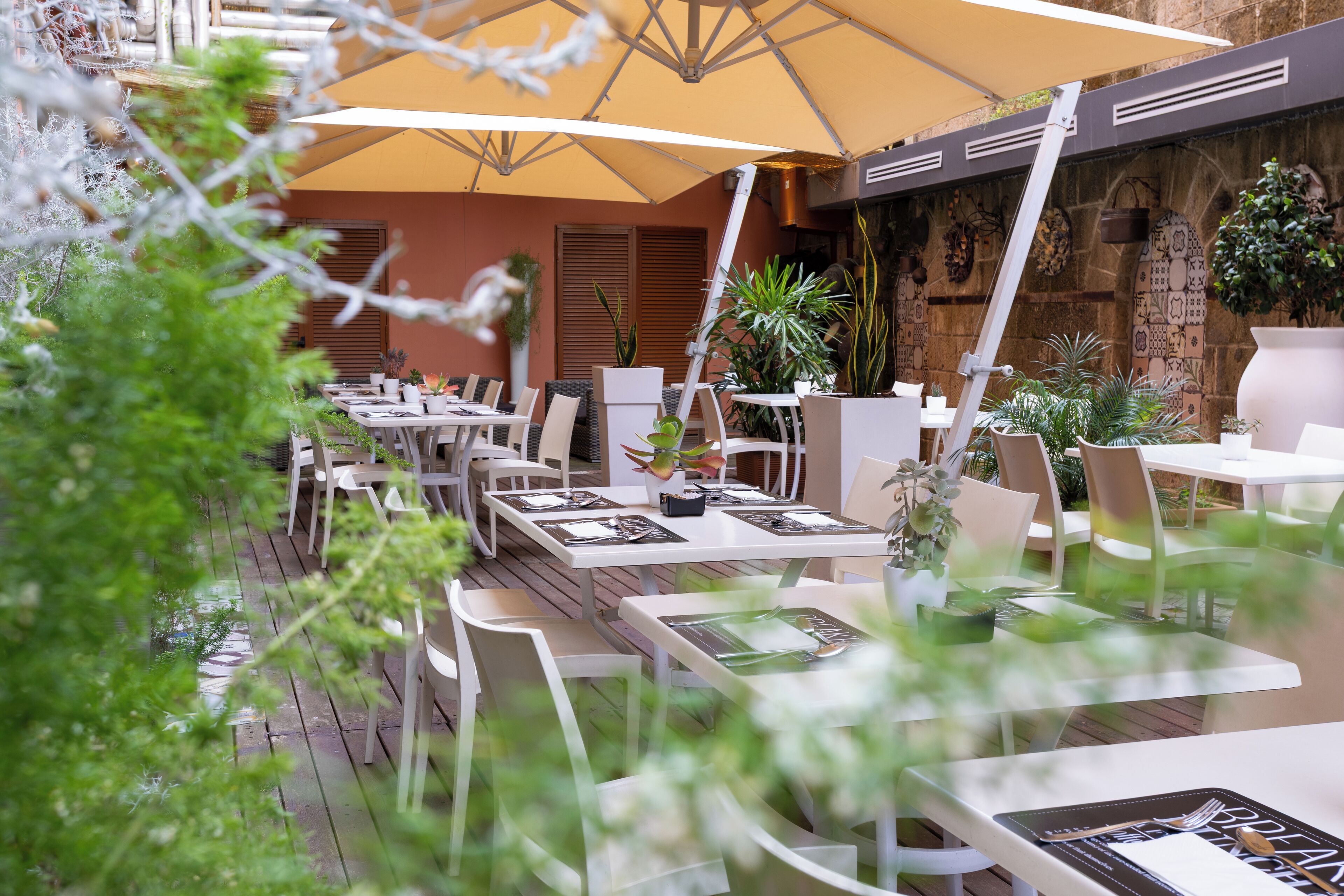 Un área de comedor al aire libre con mesas y sillas blancas, cubierta por una gran sombrilla amarilla, rodeada de plantas en macetas y un muro de piedra con detalles decorativos.