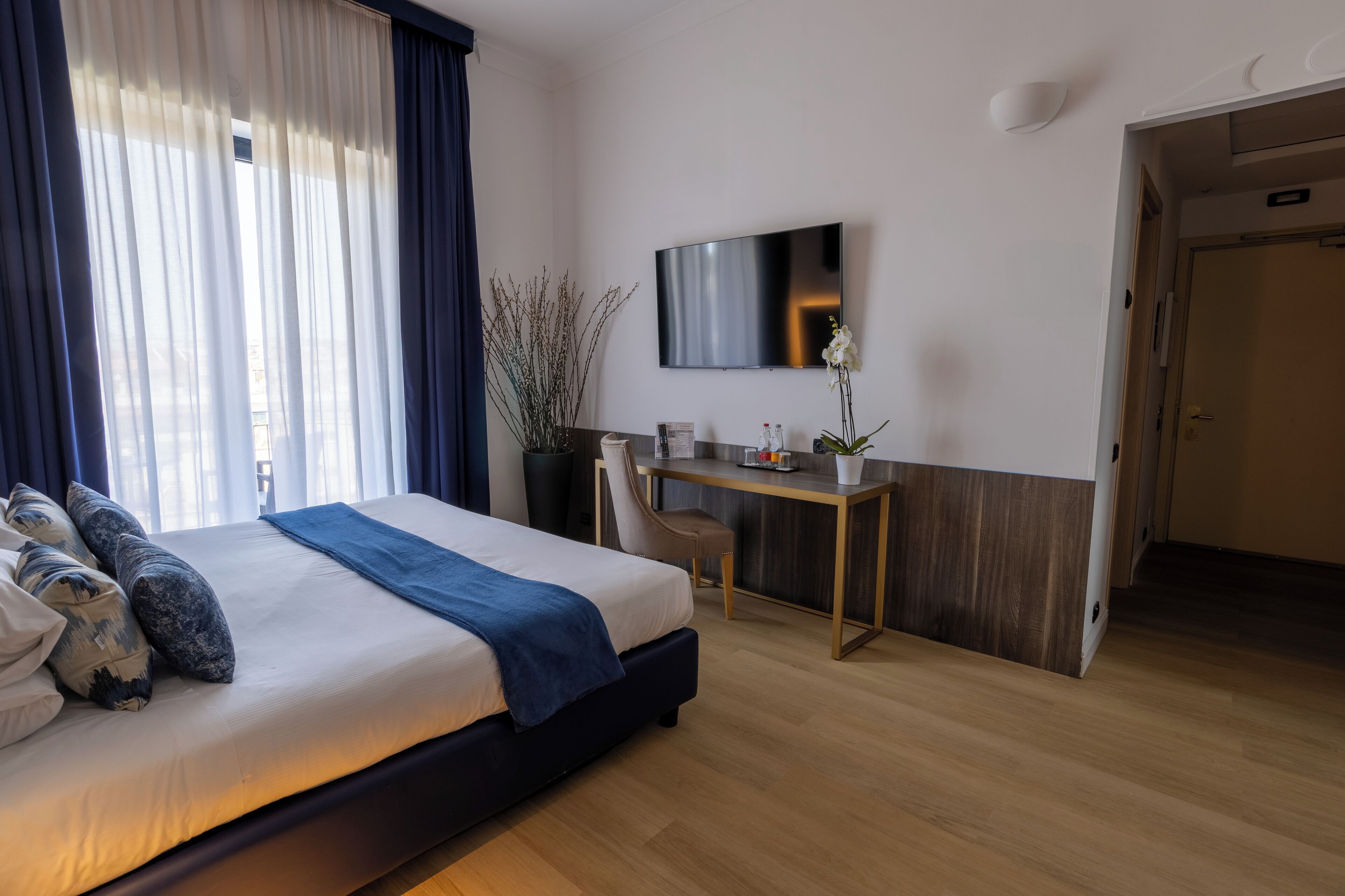 A modern hotel room with a large bed, a desk, a chair, a TV, and a door.