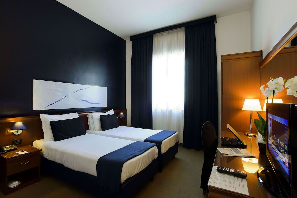 A hotel room with two beds, a desk with a computer, a TV, and a window with curtains.