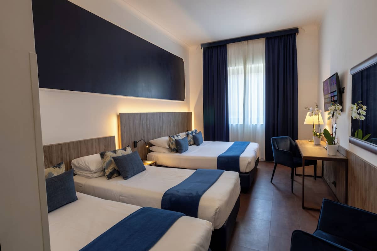 A hotel room with three beds, a desk, a chair, and a large window with curtains.