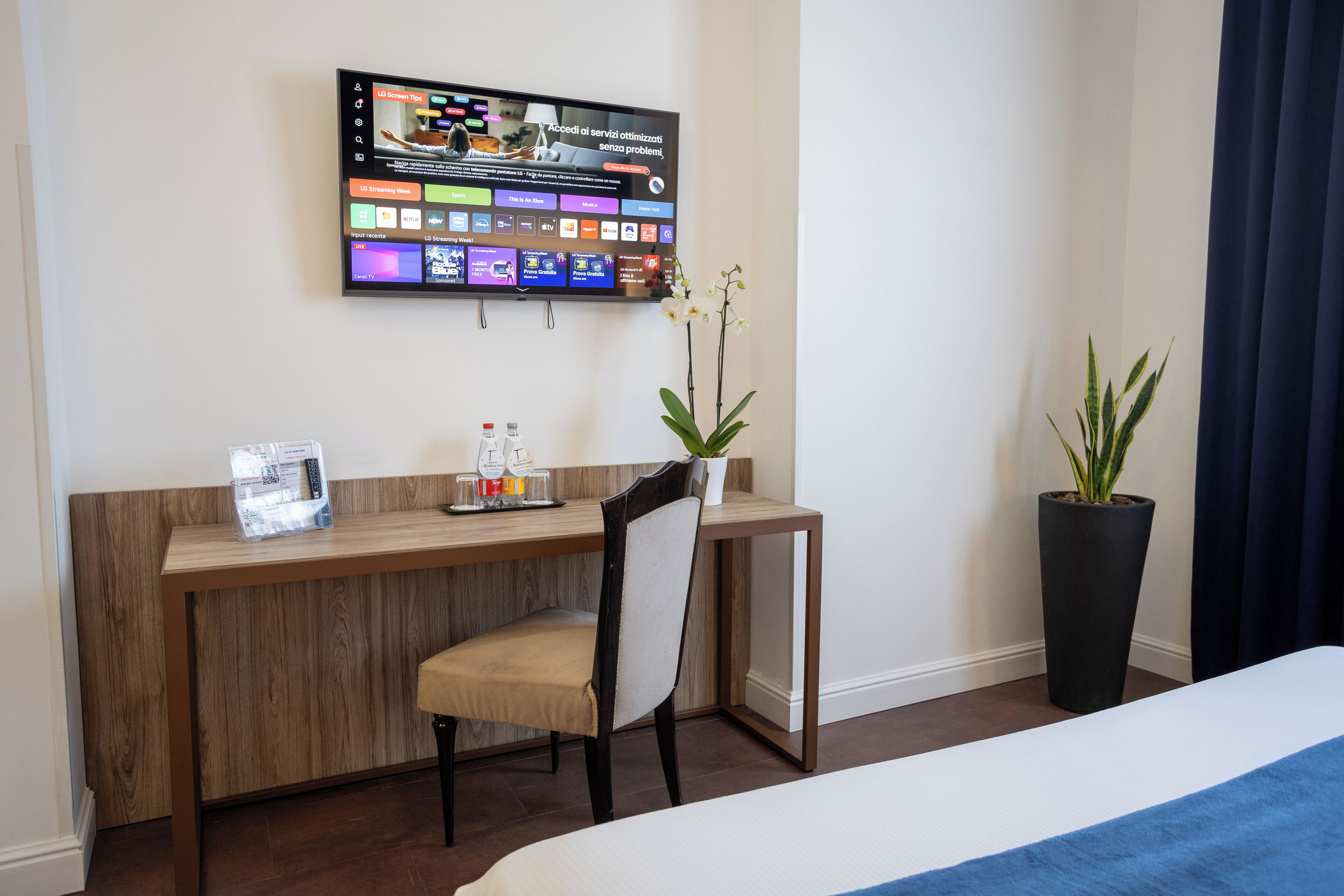 Una camera d'albergo con una scrivania in legno, una sedia, una TV a parete che mostra diversi canali e una pianta in vaso.