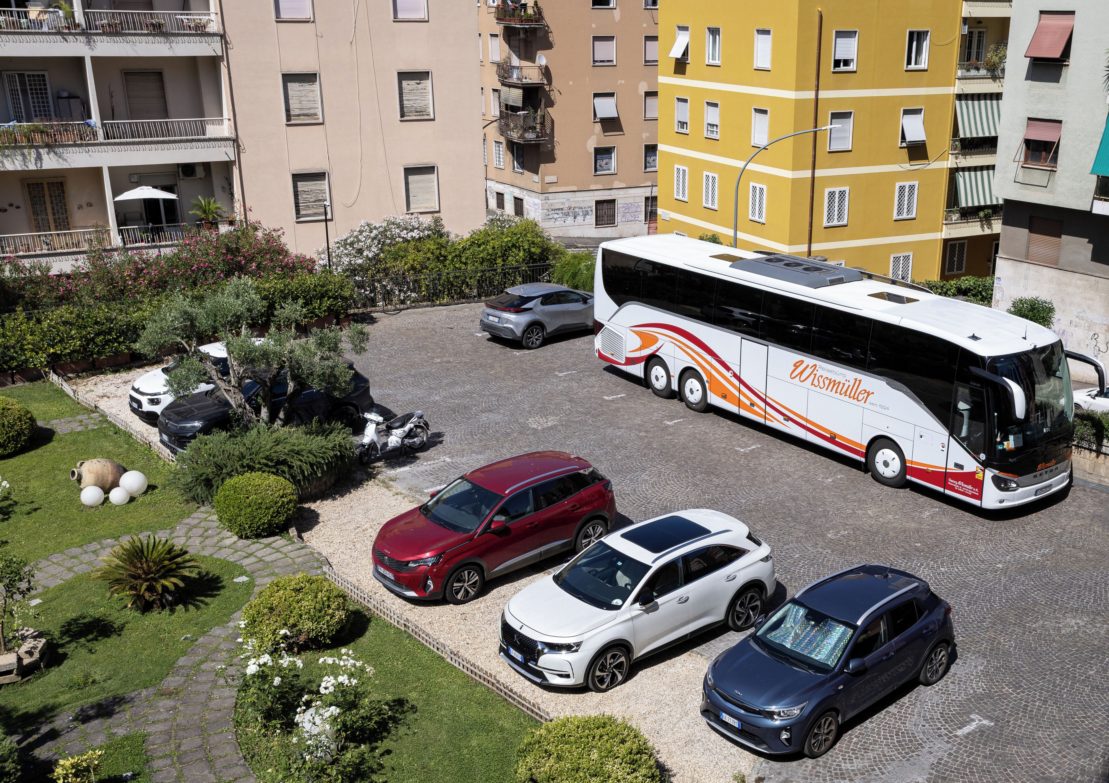 A parking area with a white bus, several cars, and greenery.