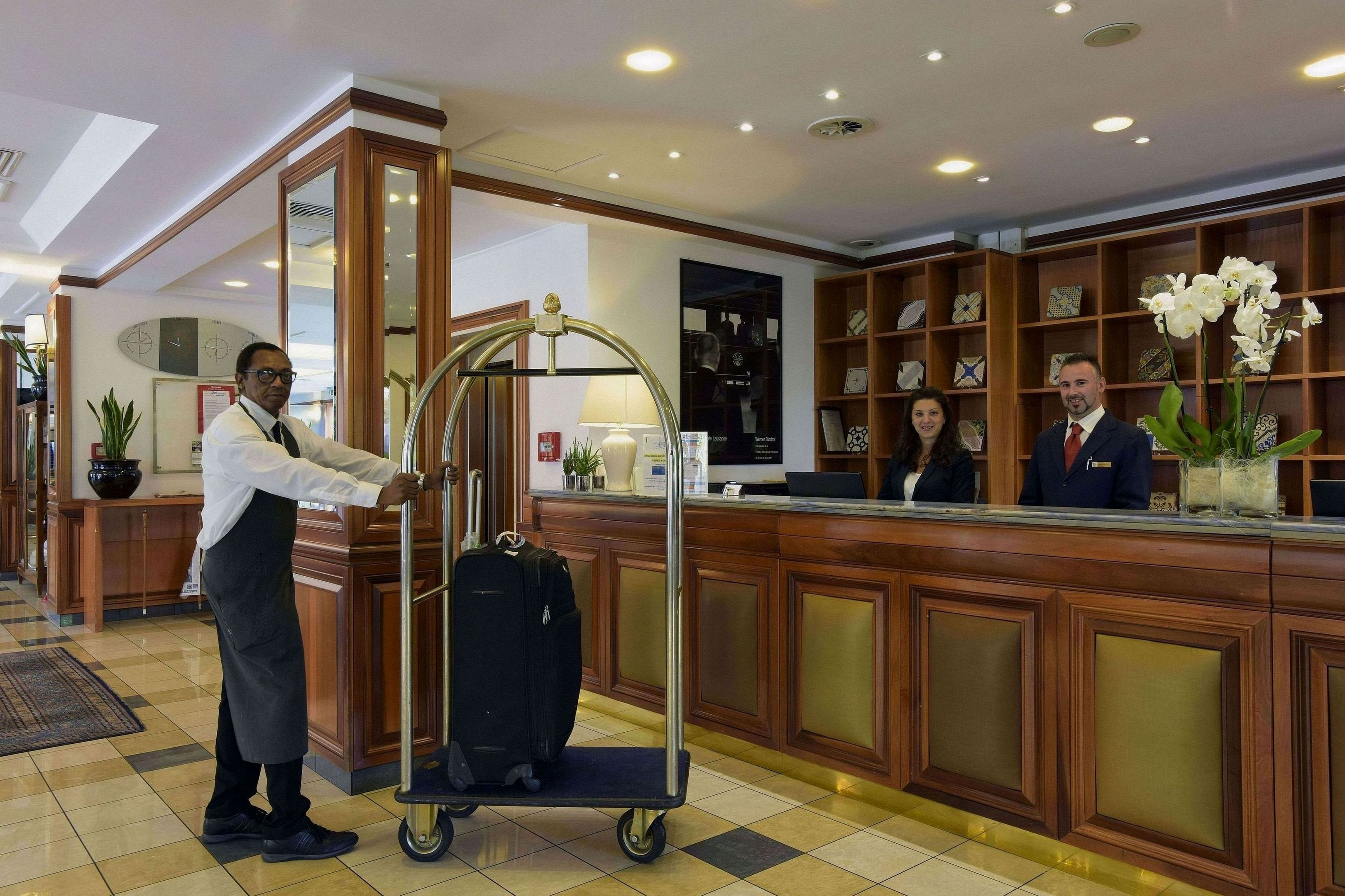 A hotel reception area with a wooden reception desk, a cart with luggage, and a reception desk with a nameplate.