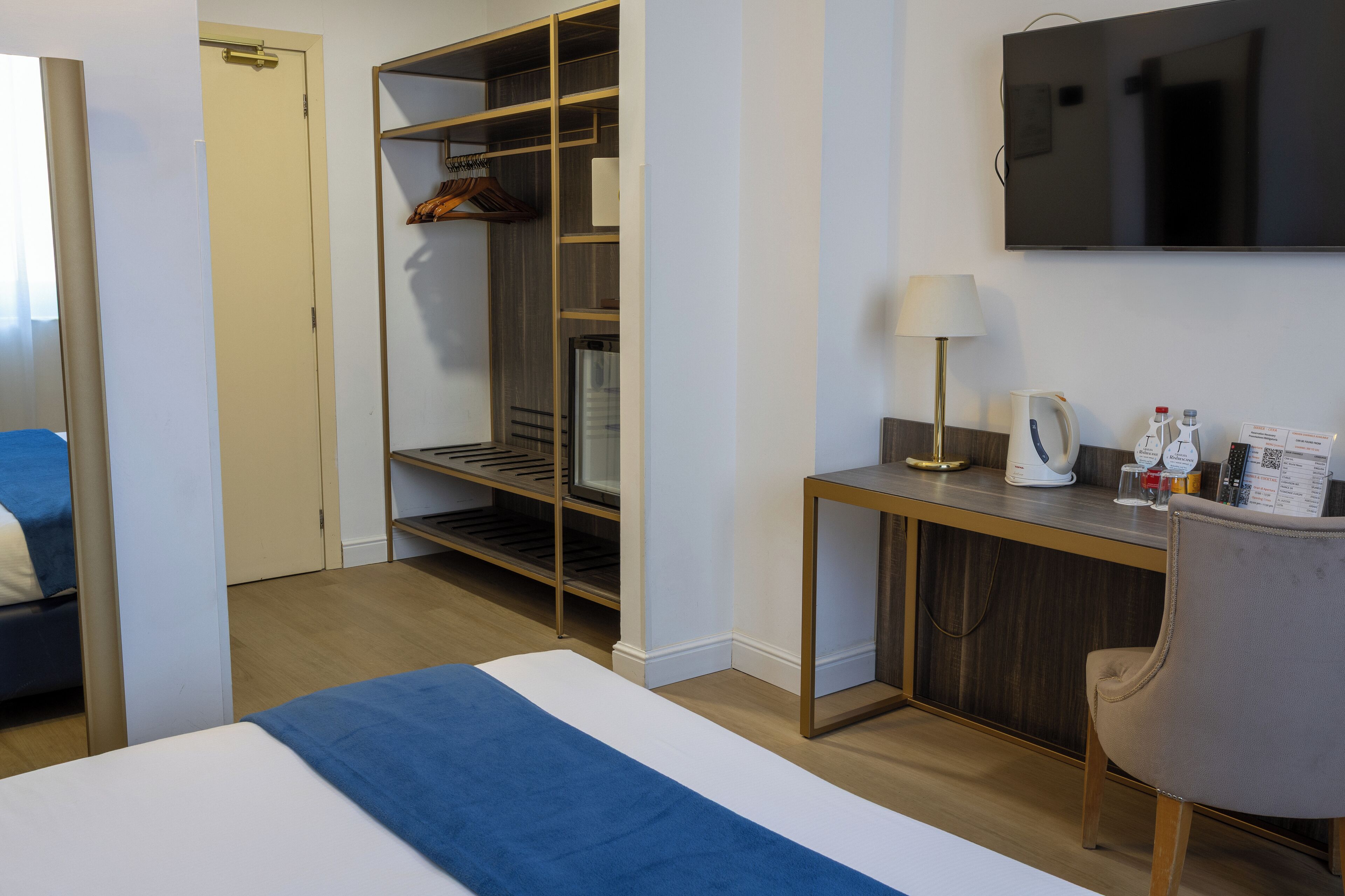 A hotel room with a bed, a desk with a lamp, a TV, a wardrobe, and a minibar.