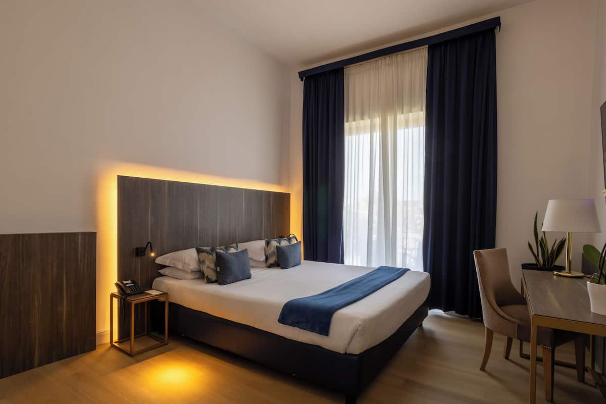 A hotel room with a large bed, bedside table with a lamp, a chair, and a small table with a plant.