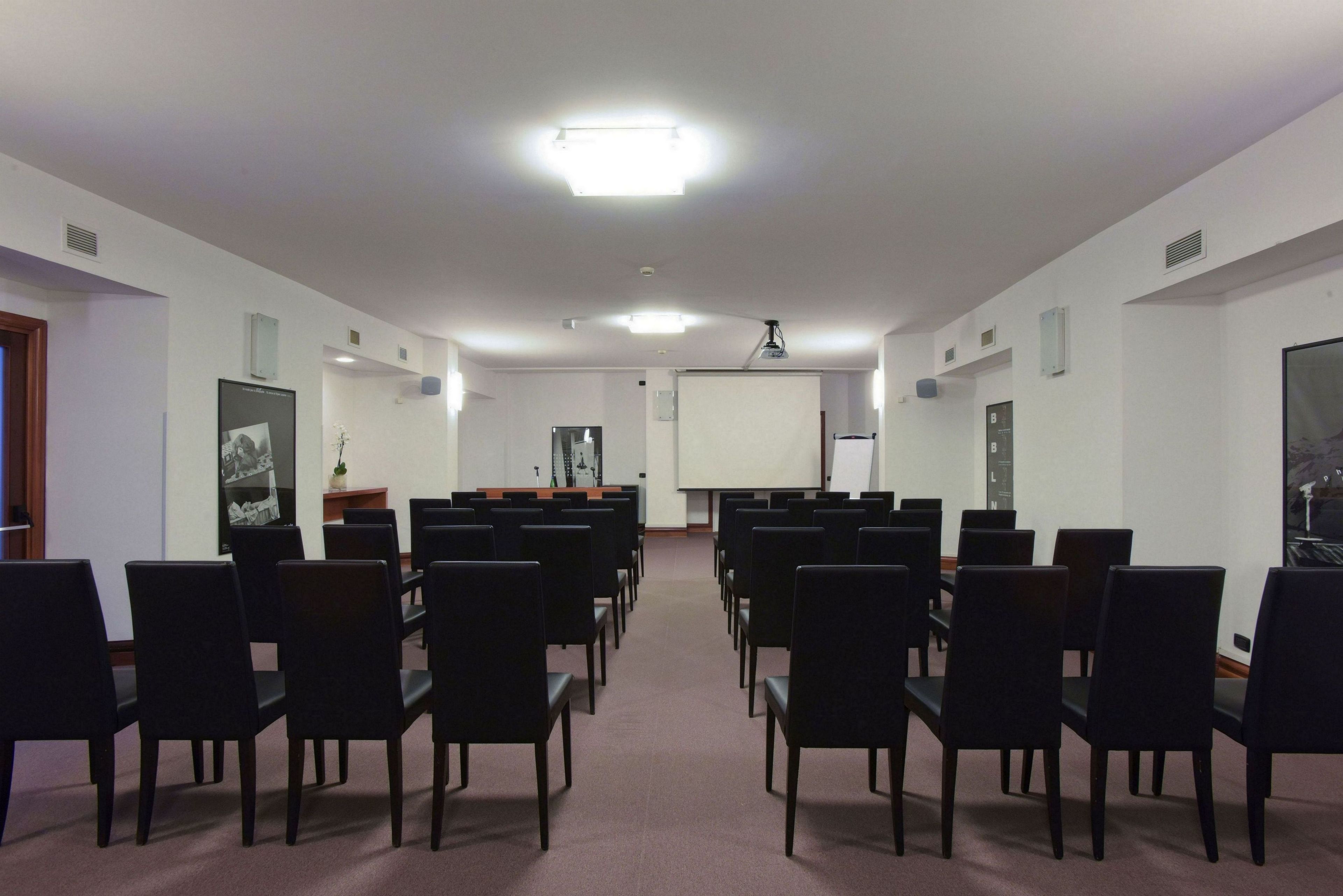Une salle de conférence avec des rangées de chaises noires, un tableau blanc et un écran de projection.