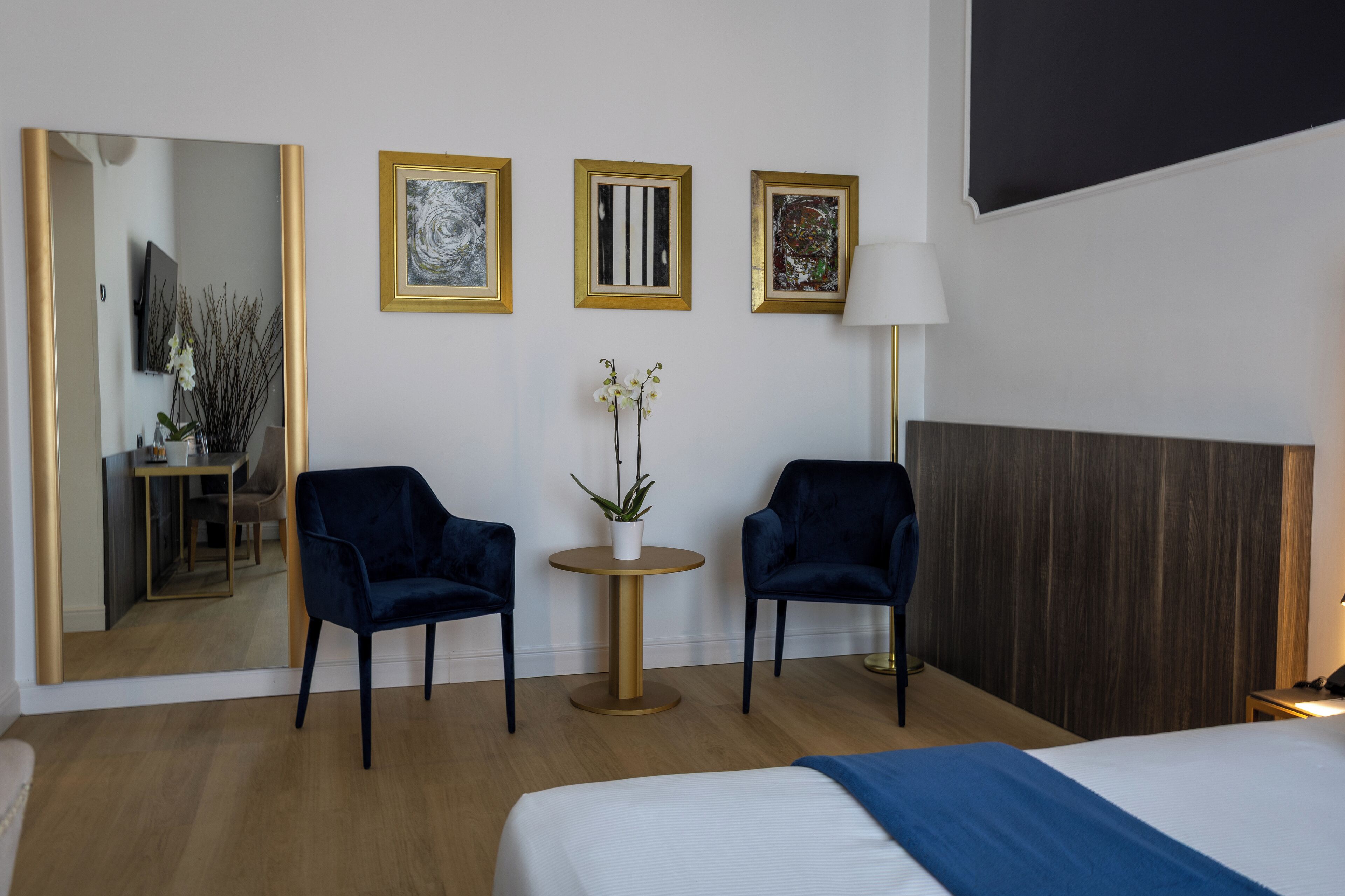 Une chambre d’hôtel avec un lit, deux fauteuils bleu foncé, une petite table ronde avec une plante, et trois œuvres d’art encadrées accrochées au mur.