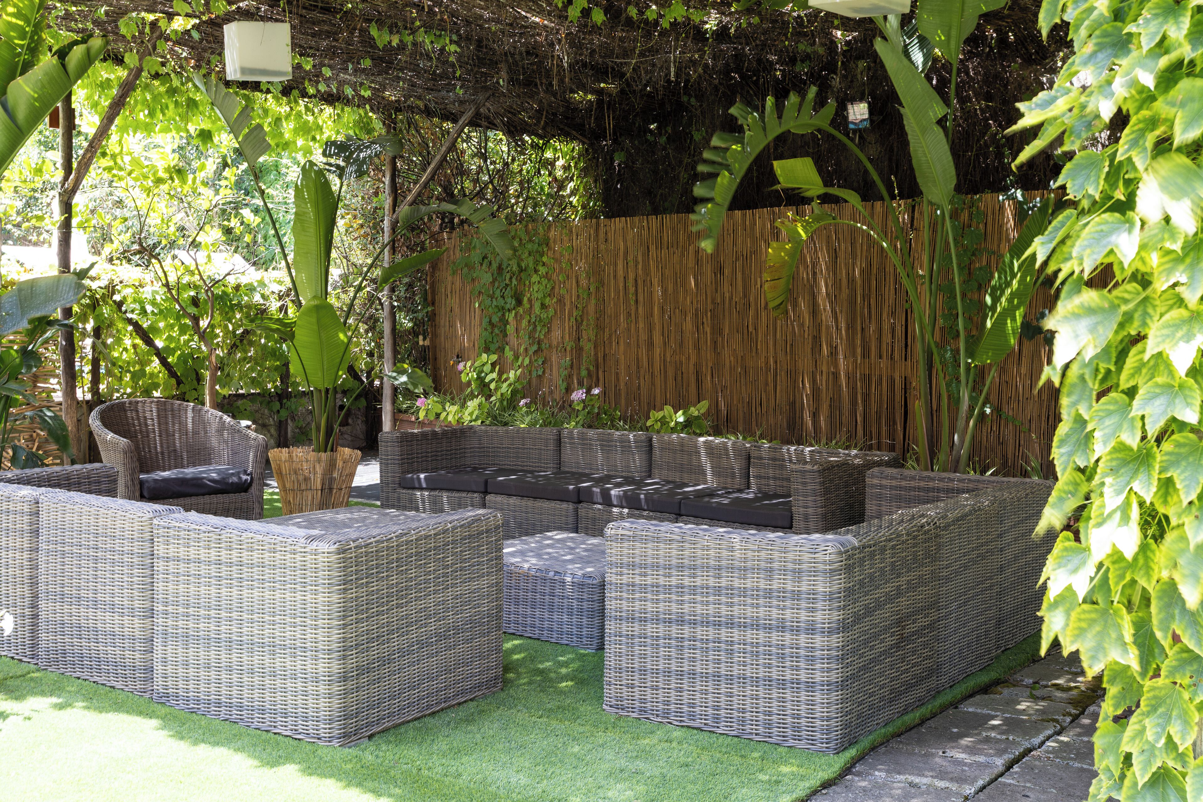 Un divano componibile in vimini con cuscini, una poltrona in vimini e un tavolino in un giardino con recinzione in legno e vegetazione.