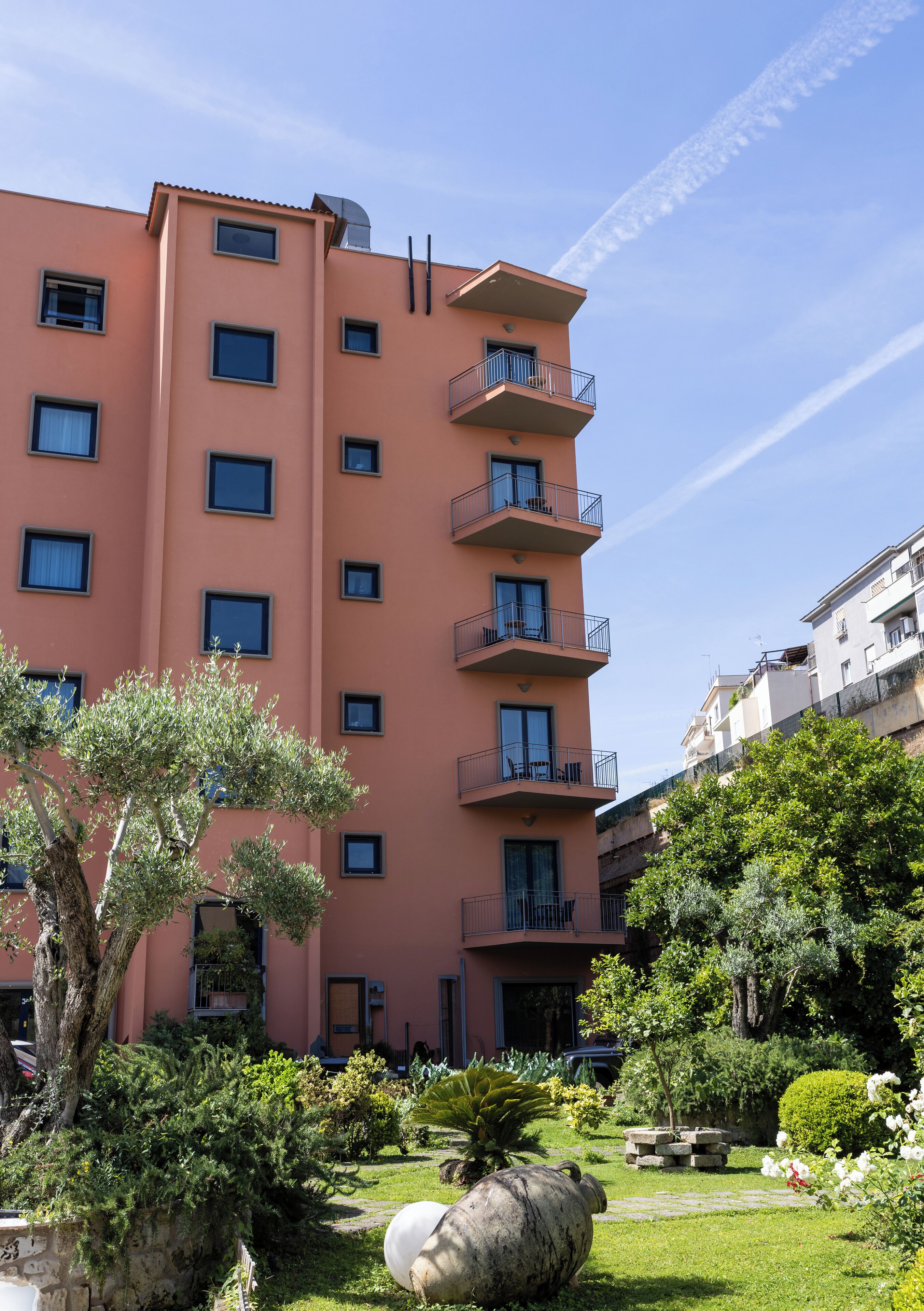 Un edificio de varios pisos con balcones, rodeado de un jardín bien cuidado con diversas plantas y una estructura de piedra.