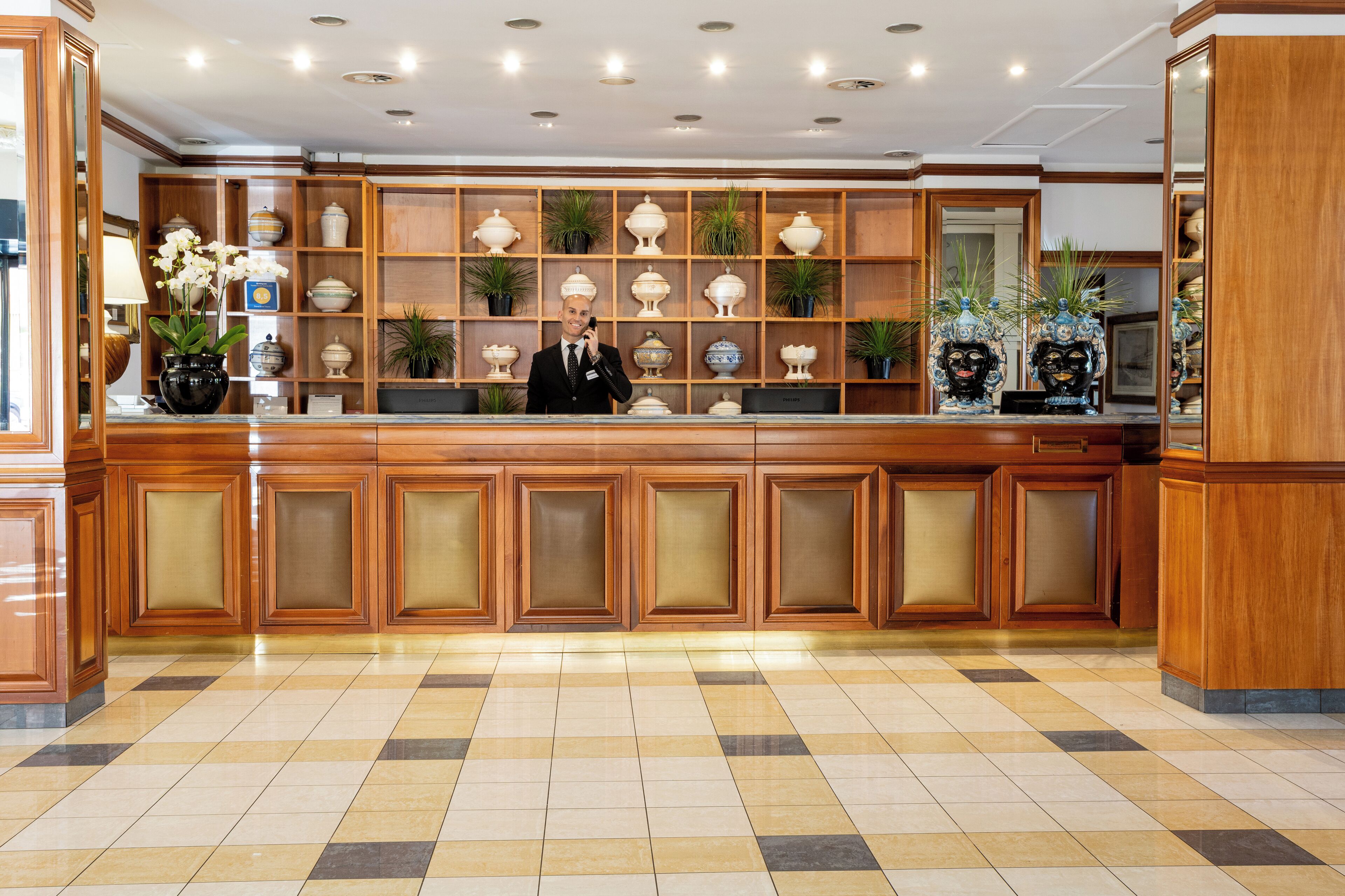 Zona de recepción de hotel con un mostrador de madera, un hombre de pie detrás y una pared decorada con jarrones y plantas.